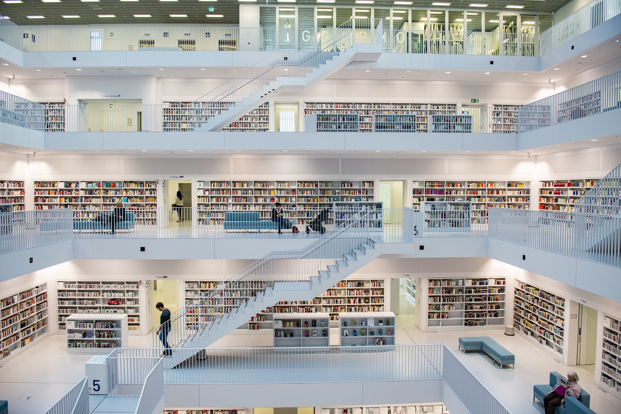 Foto da arquitetura de uma biblioteca. Aparecem 5 andares, pintados em branco, cheios de estantes com livros e pessoas circulando.