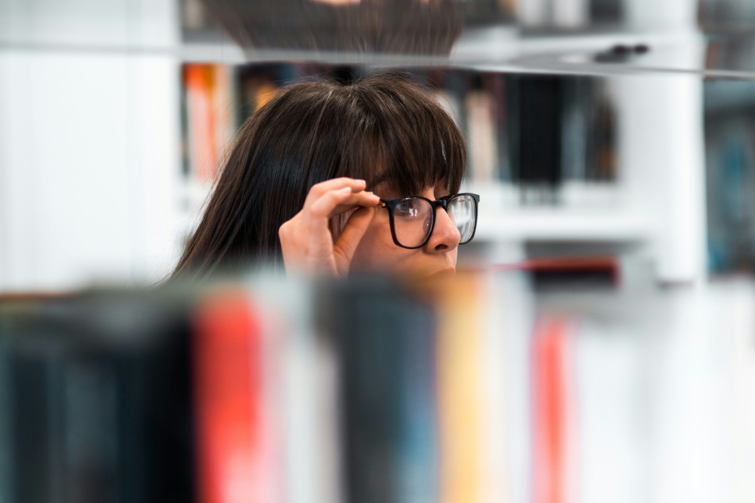 Foto de mulher usando óculos de grau enquanto olha para um acervo de livros, existe uma prateleira de livros desfocados em primeiro plano.