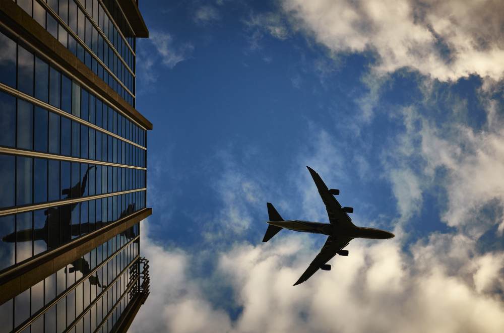 Avião, fotografado de baixo, voa por um céu de forte azul e poucas nuvens. À esquerda um edifício comercial espelha a aeronave.