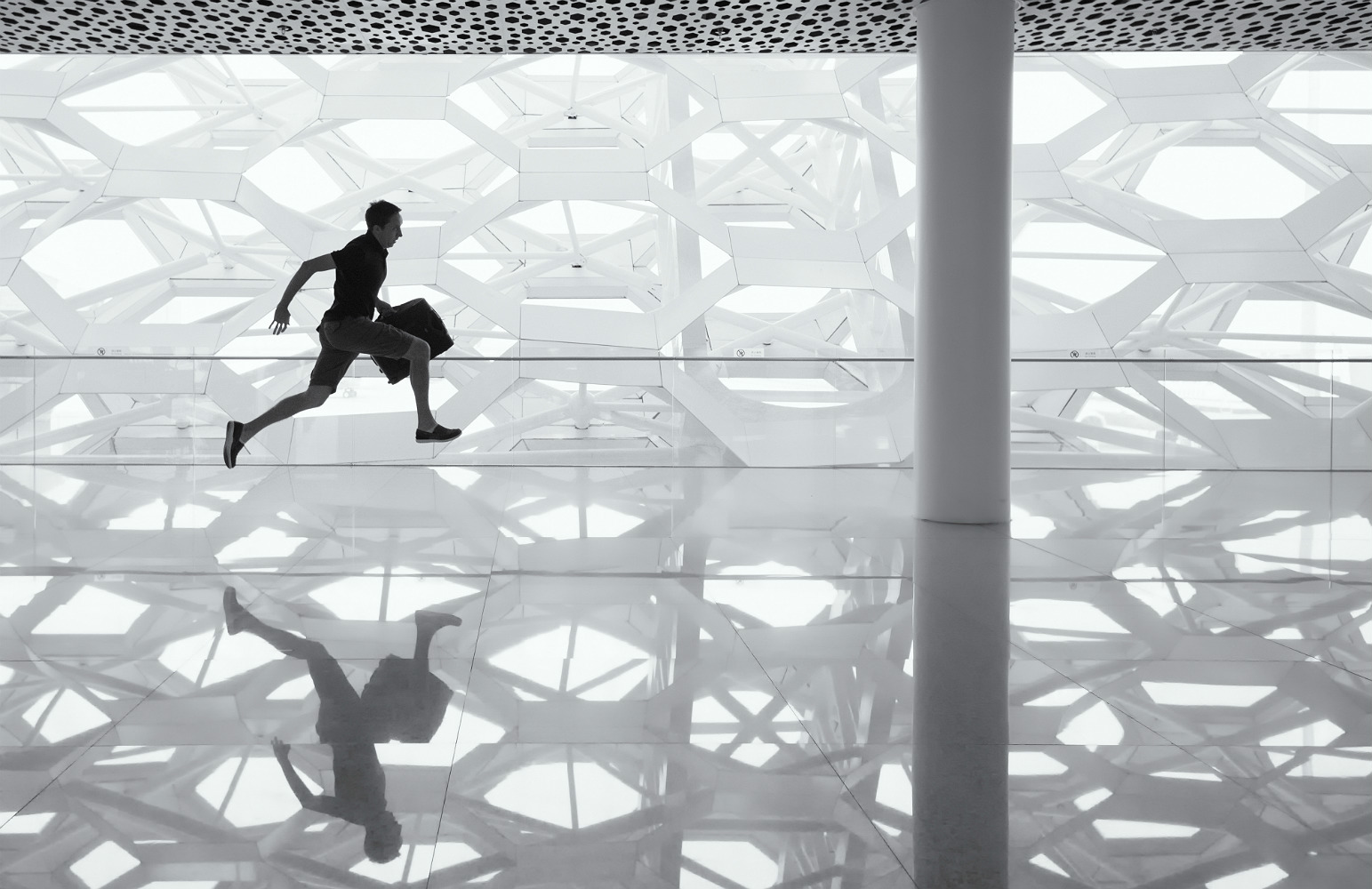 Imagem em preto e branco de um homem saltando apressado em um saguão. O homem porta uma mala e o chão lustrado reflete seu salto.