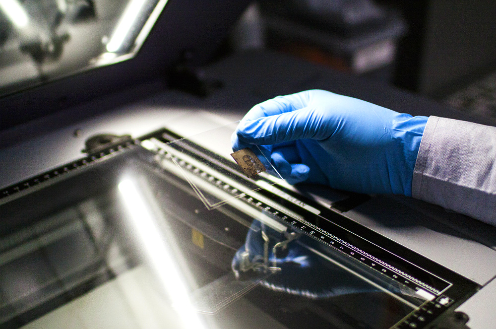 Uma mão envolta em uma luva azul de látex colocando uma foto antiga sobre um aparelho de scanner.
