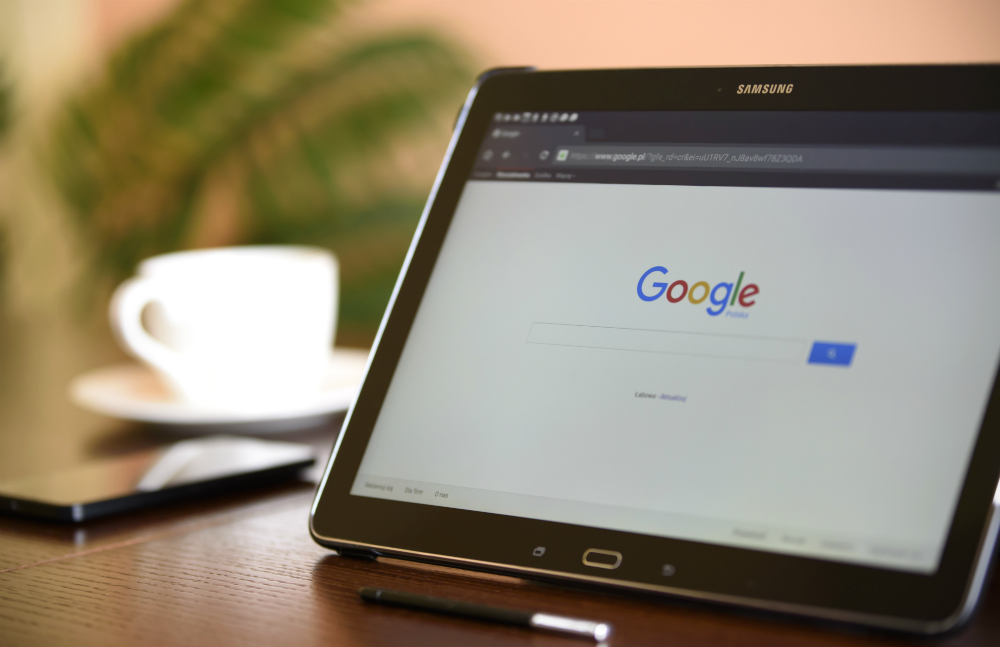 No primeiro plano, sobre uma superfície de madeira escura, um tablet inclinado para cima à direita mostra a tela inicial da busca do Google. Ao fundo, desfocados, uma xícara branca, folhas de uma árvore ornamental e uma parede rosada.