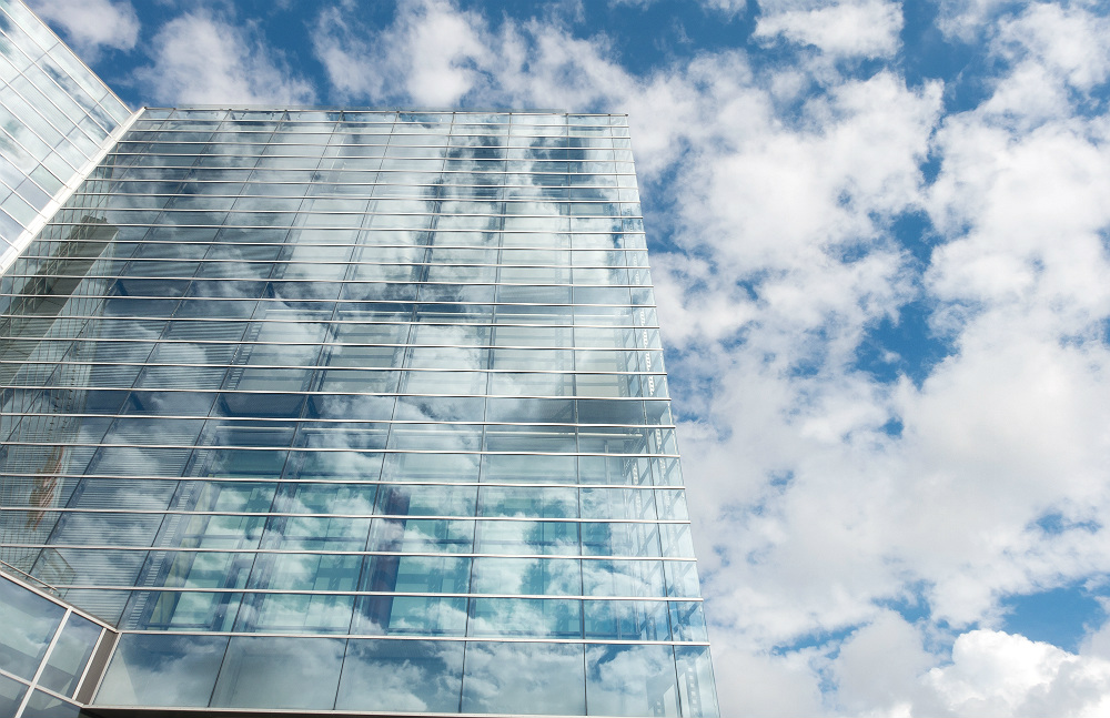 Foto de um prédio com a fachada de vidro, que reflete as nuvens e a cor do céu, no fundo do prédio está o céu.
