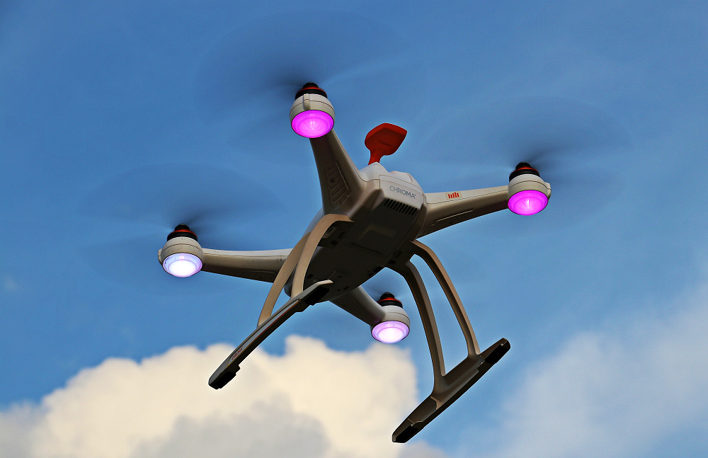 Close em um drone visto de baixo, voando por um céu azul com poucas nuvens, ascendendo luzes rosadas e brancas.