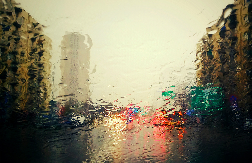 A visão de uma rua em fim de tarde, com placas, faróis vermelhos e prédios, embaçada pela chuva sobre uma superfície transparente.