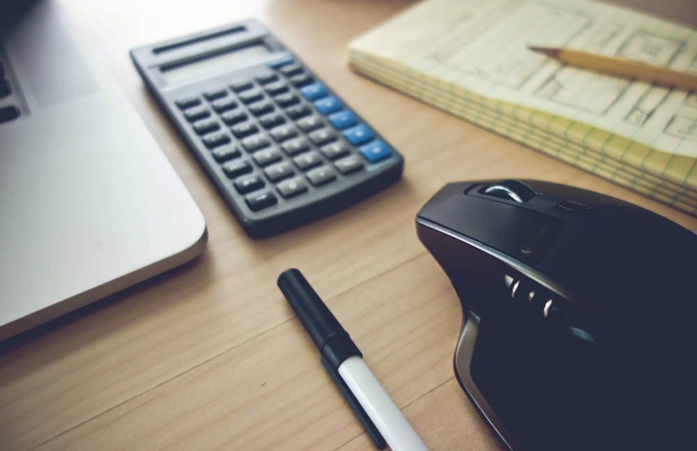 Close em uma calculadora preta sobre uma superfície de madeira, cercada por uma caneta, um laptop, um mouse e um bloco de papel com um lápis.