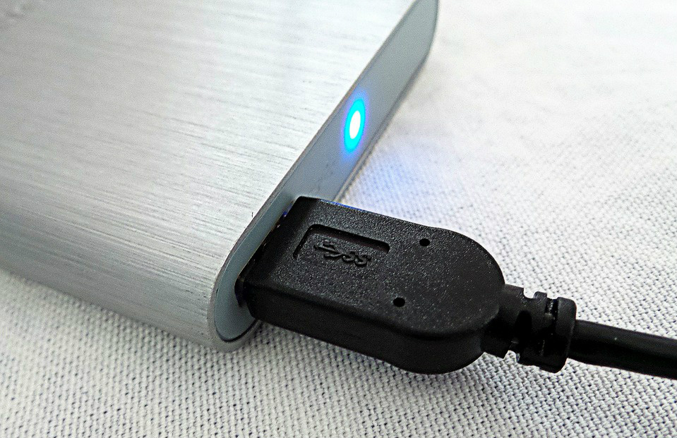 HD externo prateado com uma luz azul acesa, e com cabo USB conectado, sobre uma superfície da mesma cor.