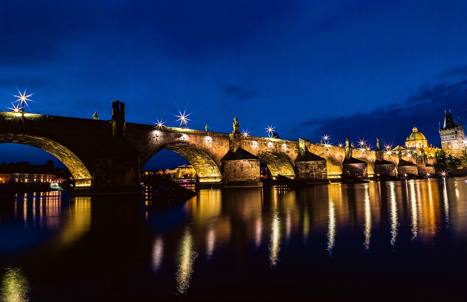 A icônica Ponte Carlos, em Praga, na Tchéquia, fotografada à noite, com sua iluminação refletindo nas águas do rio Moldava.