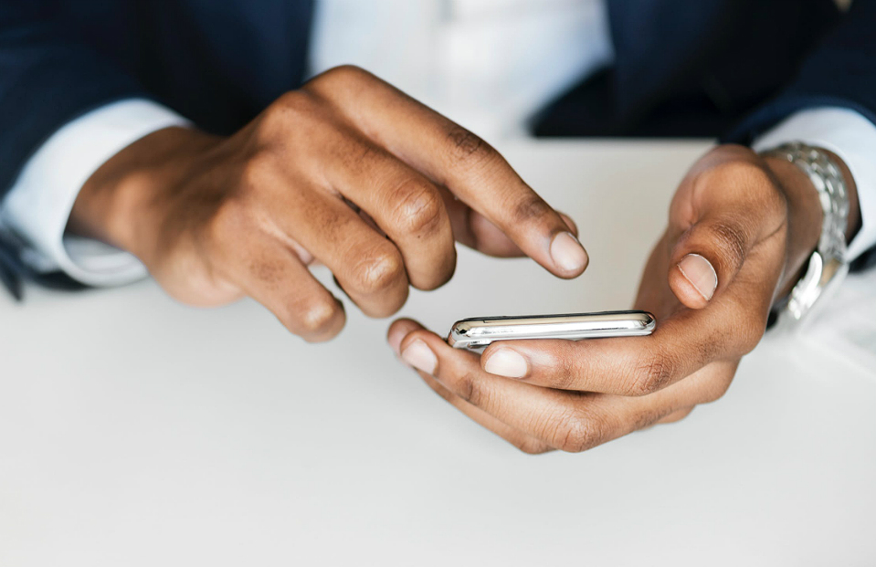 Close nas mãos de uma pessoa vestindo paletó. Uma mão segura um smartphone e a outra toca na tela do aparelho com o dedo indicador.