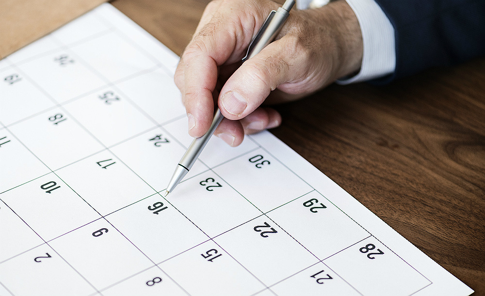 Foto de uma mão segurando uma caneta apontando para um dia específico de um calendário.