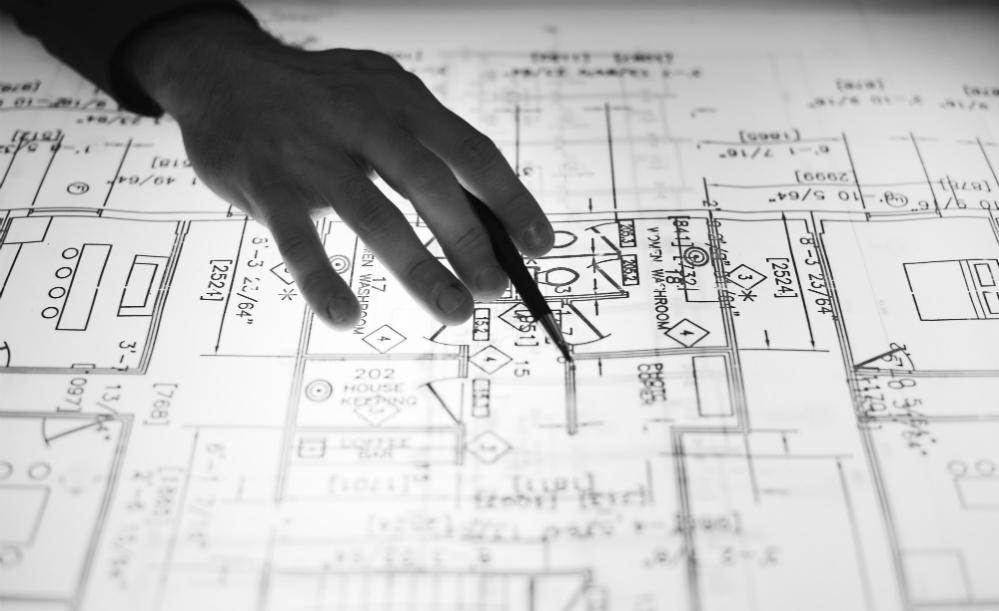 Fotografia em preto e branco mostra uma mão manipulando uma caneta ou lapiseira sobre uma planta de construção, por sua vez colocada sobre uma mesa de luz.