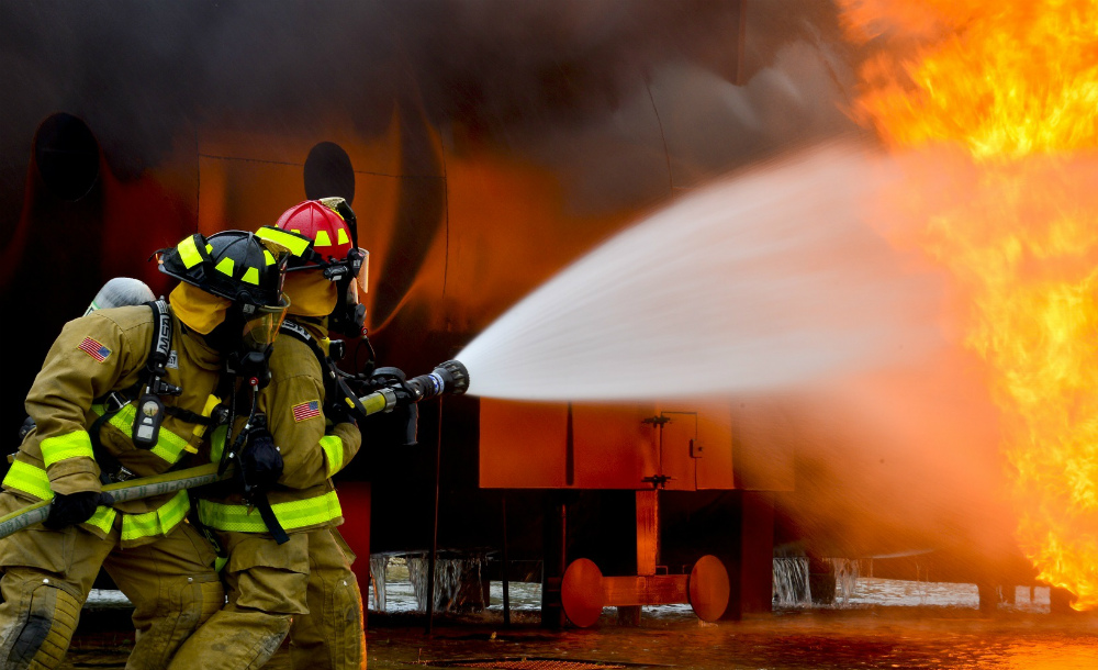 À esquerda, dois bombeiros uniformizados da cabeça aos pés miram um potente jato de água para as chamas no lado direito da imagem.
