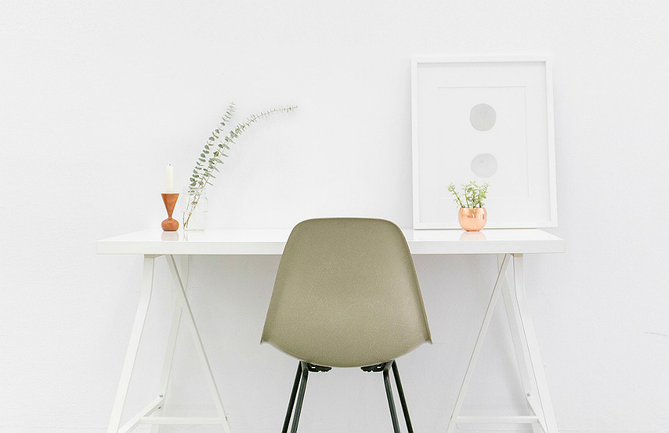 Foto de uma sala branca com uma mesa também branca, organizada com quadro, vaso de planta e suporte de vela, e uma cadeira acinzentada.