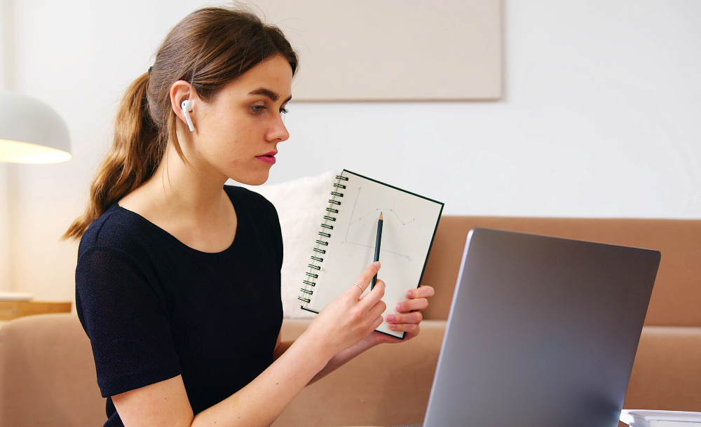 Foto de mulher em videochamada, com fones de ouvido, de frente para um notebook, ela está segurando um caderno virado para a tela, apontando para um gráfico desenhado nele com um lápis.