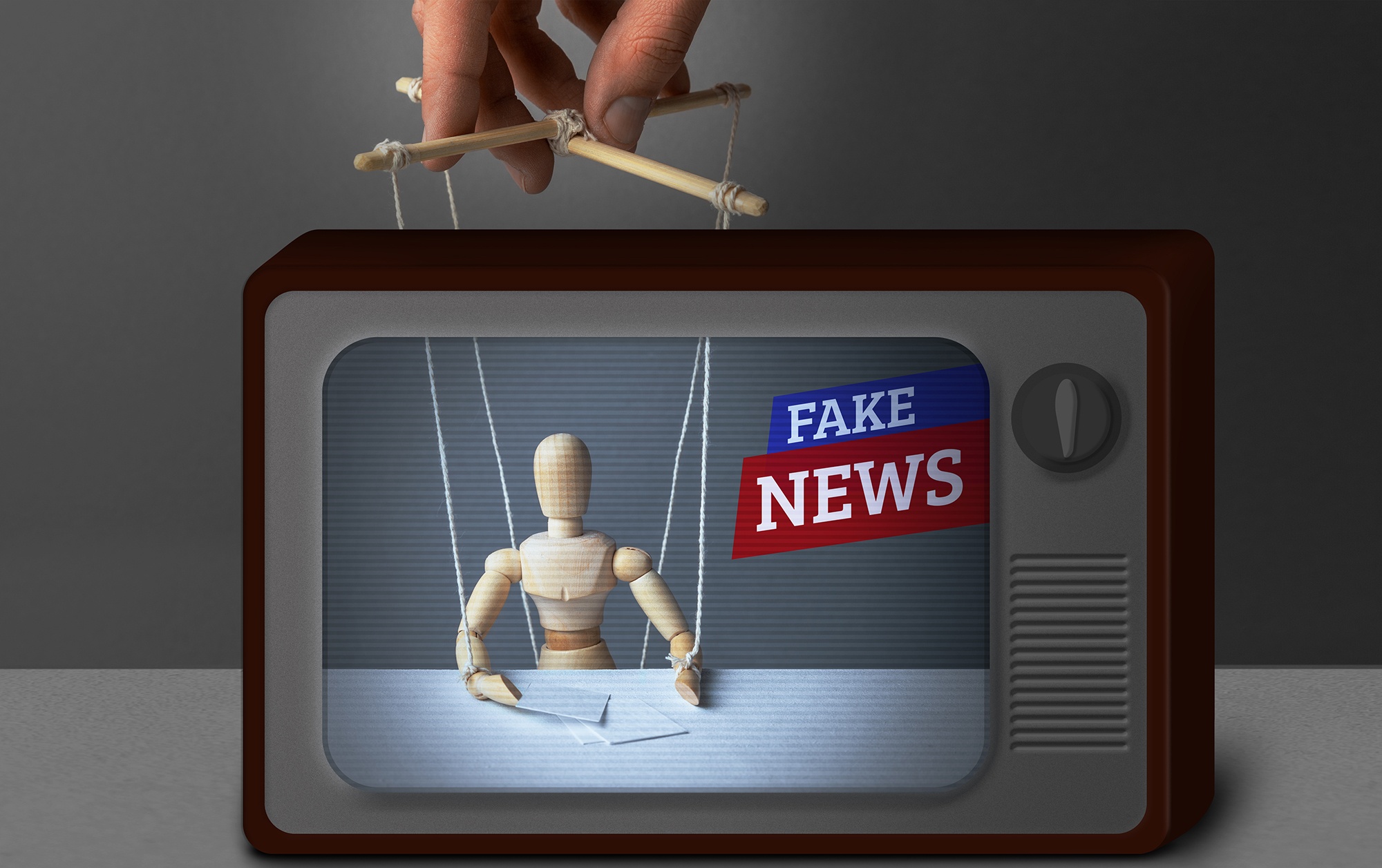 A foto mostra a tela de uma televisão antiga que apresenta a imagem de um boneco de madeira articulado com cordas amarrada (marionete) e os dizeres "Fake News".