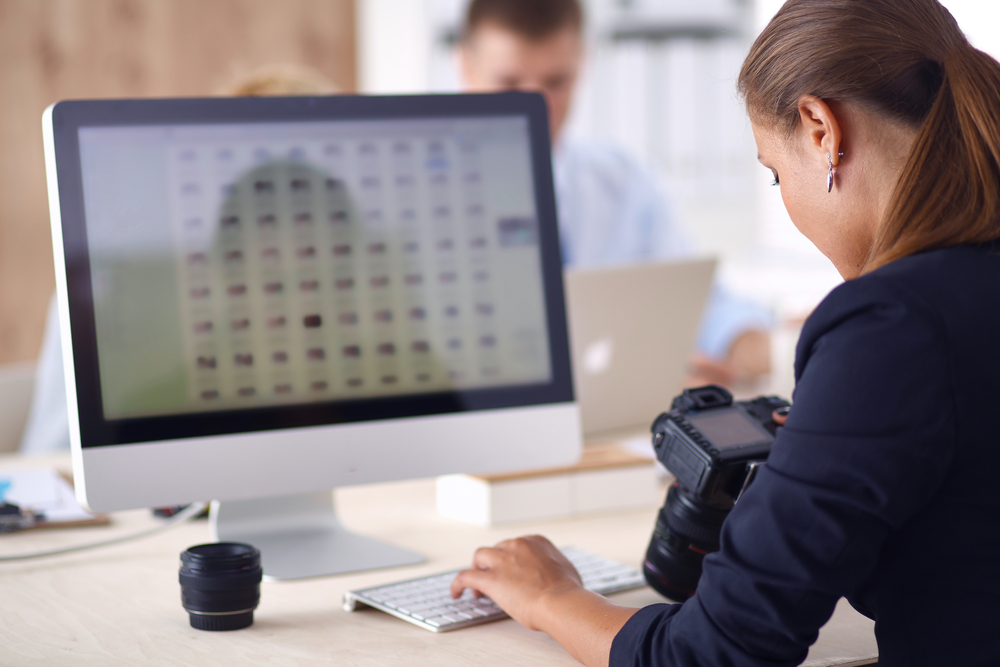 A foto mostra uma mulher de frente para um computador enquanto segura e olha para uma câmera fotográfica.