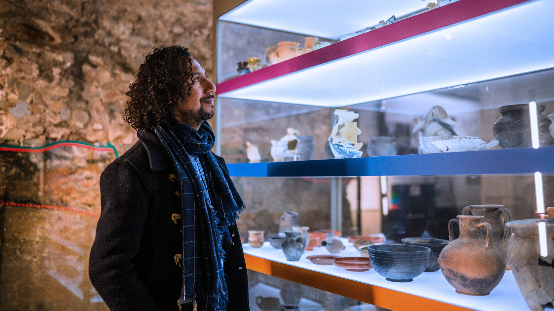 Homem olha para estante com vasos antigos protegidos por vidro em uma exposição.